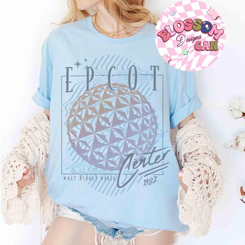 May include: Light blue t-shirt featuring a graphic of the EPCOT Center sphere with the text "EPCOT Center Walt Disney World 1982". The design is in shades of gray and purple. The shirt has rolled-up sleeves.