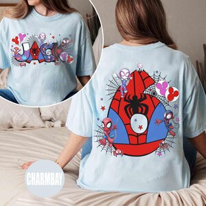 Pu&ograve; includere: T-shirt azzurra con un design colorato di Spider-Man. La parte anteriore mostra il nome "JAKE" con ragnatele e grafiche dei personaggi. Il retro presenta un grande emblema di Spider-Man con vari personaggi e dettagli di ragnatele. Il marchio "CHARMBAY" &egrave; visibile.