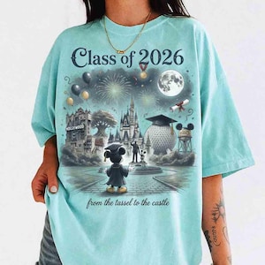 May include: A light blue t-shirt with a graphic featuring Mickey Mouse in a graduation cap, the text "Class of 2026", and the phrase "from the tassel to the castle". The design includes fireworks, a castle, and a full moon.
