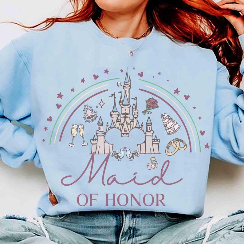 May include: Light blue sweatshirt with a graphic featuring a castle, rainbow, and the words "Maid of Honor." The design includes champagne glasses, a cake, rings, and Mickey Mouse silhouettes. A perfect gift for a bachelorette party or bridal shower.