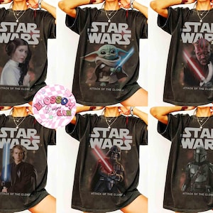 May include: A collection of dark gray t-shirts featuring Star Wars characters. Each shirt displays the Star Wars logo and the text "ATTACK OF THE CLONE". The shirts showcase images of Leia, Yoda, Darth Maul, Anakin, Darth Vader, and The Mandalorian.