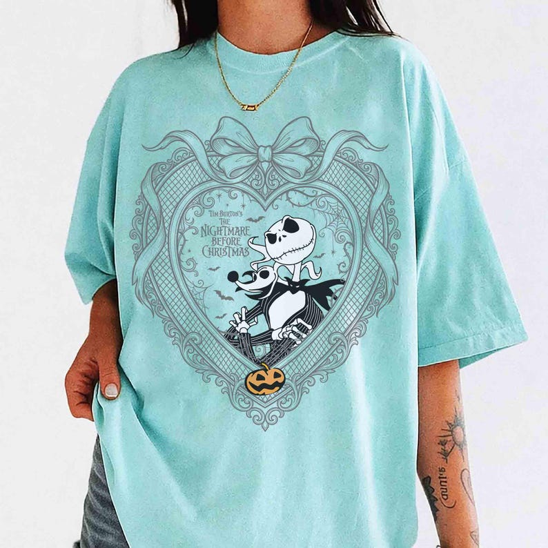 May include: A light blue t-shirt featuring a heart-shaped design with characters from "The Nightmare Before Christmas." The design includes Jack Skellington, Zero, and a pumpkin. The shirt has short sleeves and a relaxed fit.