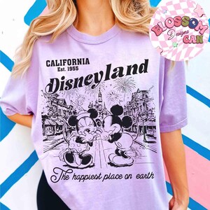 May include: A lilac-colored t-shirt featuring a black and white graphic of Mickey and Minnie Mouse in front of Disneyland. The text reads "CALIFORNIA Est. 1955 Disneyland" and "The happiest place on earth."
