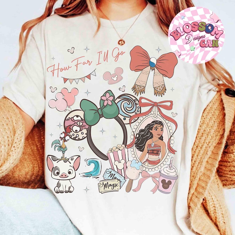 May include: Cream-colored t-shirt with a colorful graphic design featuring the text "How Far I'll Go" and various cartoon elements. The design includes a portrait of a character, bows, and other themed illustrations.
