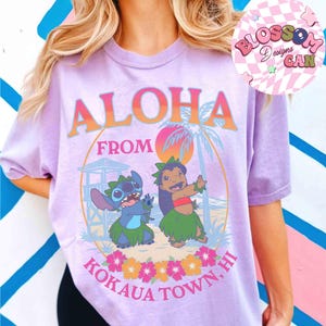 May include: A lilac t-shirt with the words "ALOHA FROM KOKAUA TOWN, HI" in orange and blue. The shirt features a graphic of Stitch and Lilo in front of a beach scene with palm trees and flowers. The shirt has short sleeves.