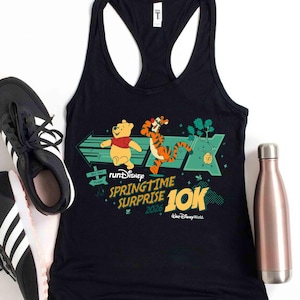 Könnte beinhalten: Schwarzes Tanktop mit einem Design von Winnie Puuh und Tigger, mit dem Text "runDisney Springtime Surprise 2026 10K". Das Design umfasst grüne Pfeile und Akzente, mit dem Walt Disney World Logo.