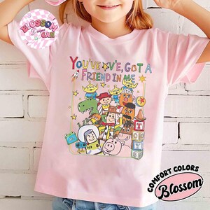 May include: A light pink t-shirt featuring a colorful graphic of characters and the text "YOU'VE GOT A FRIEND IN ME". The design includes a rocket, stars, and a variety of cartoon figures. The shirt also has the text "COMFORT COLORS" and "Blossom".