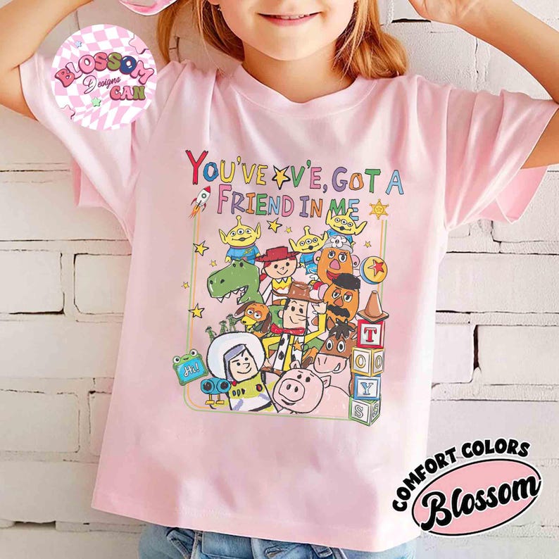 May include: A light pink t-shirt featuring a colorful graphic of characters and the text "YOU'VE GOT A FRIEND IN ME". The design includes a rocket, stars, and a variety of cartoon figures. The shirt also has the text "COMFORT COLORS" and "Blossom".