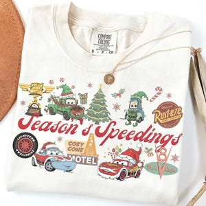 May include: Cream-colored t-shirt featuring a festive graphic with Cars characters in Santa hats and holiday decorations. The text "Season's Speedings" is in red script. A gold necklace with a pendant is draped on the shirt.
