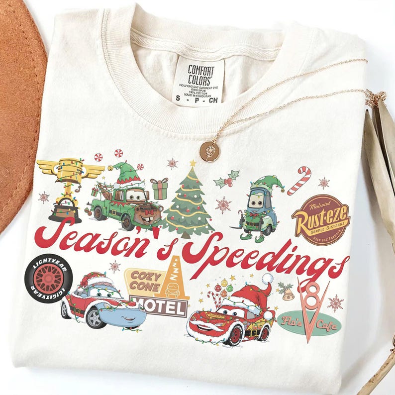 May include: Cream-colored t-shirt featuring a festive graphic with Cars characters in Santa hats and holiday decorations. The text "Season's Speedings" is in red script. A gold necklace with a pendant is draped on the shirt.
