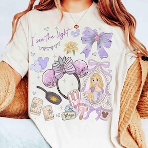 Op de afbeelding: Een crèmekleurig t-shirt met een grafische print met de tekst I see the light, een portret van een blond personage in een paarse jurk, een koekenpan, oren met een strik, een grote paarse strik, lantaarns en diverse kleine iconen.