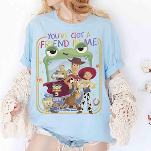 May include: A light blue t-shirt featuring a graphic print of Toy Story 5 characters including Woody, Buzz Lightyear, Jessie, and Bullseye. The text reads "YOU'VE GOT A FRIEND IN ME" and "Disney PIXAR TOY STORY 5 IN THEATERS JUNE 19" inside a frame.