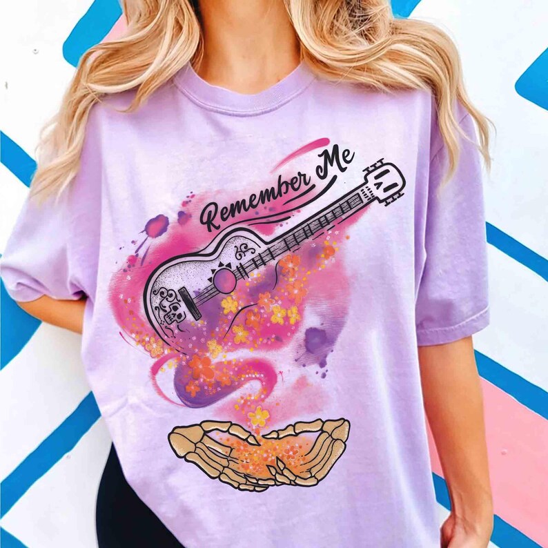 Puede incluir: Camiseta lila claro con un dise&ntilde;o de guitarra y el texto "Remember Me". La guitarra est&aacute; decorada con motivos florales y de calaveras, con un efecto acuarela colorido. Una mano de esqueleto est&aacute; en la parte inferior.