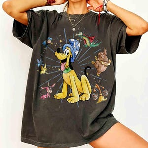 Może przedstawiać: Ciemnoszara koszulka z kreskówkową ilustracją Plutona w czarodziejskim kapeluszu, otoczonego innymi postaciami z kreskówek. Projekt zawiera żółtego psa, zielonego smoka i różowego jednorożca. Koszulka ma wygląd vintage.