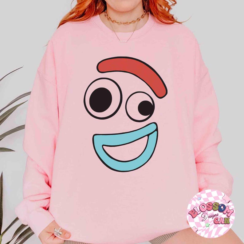 May include: A light pink sweatshirt featuring a cartoon face with black eyes, a red eyebrow, and a blue mouth. The sweatshirt has a crew neck and long sleeves. A small, circular logo is visible on the lower right.