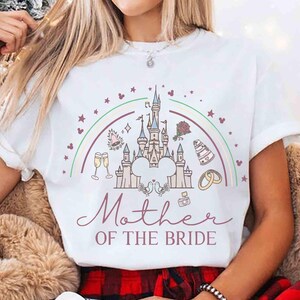 May include: White t-shirt featuring a castle design with a rainbow, stars, and wedding-themed illustrations. The text "Mother of the Bride" is written in a script font. The shirt is a short-sleeved crew neck.