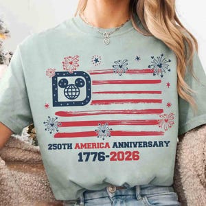 May include: A light green t-shirt featuring a stylized American flag design with red stripes and a blue emblem. The emblem incorporates a Mickey Mouse head and a globe. Text reads "250TH AMERICA ANNIVERSARY 1776-2026".