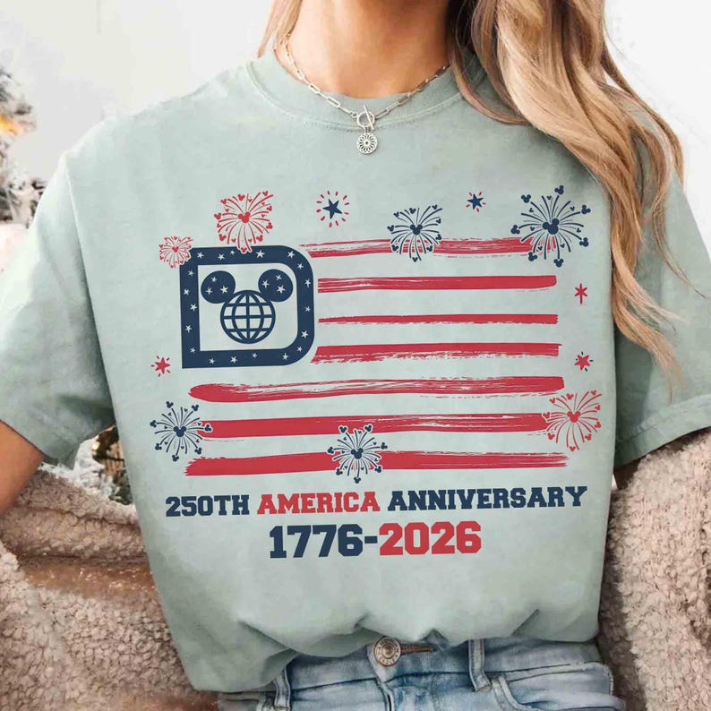 May include: A light green t-shirt featuring a stylized American flag design with red stripes and a blue emblem. The emblem incorporates a Mickey Mouse head and a globe. Text reads "250TH AMERICA ANNIVERSARY 1776-2026".