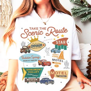 May include: White t-shirt featuring a colorful graphic design with the text "Take the Scenic Route." The design includes illustrations of cars, cacti, and various signs, including "Radiator Springs" and "Motel No Vacancy."