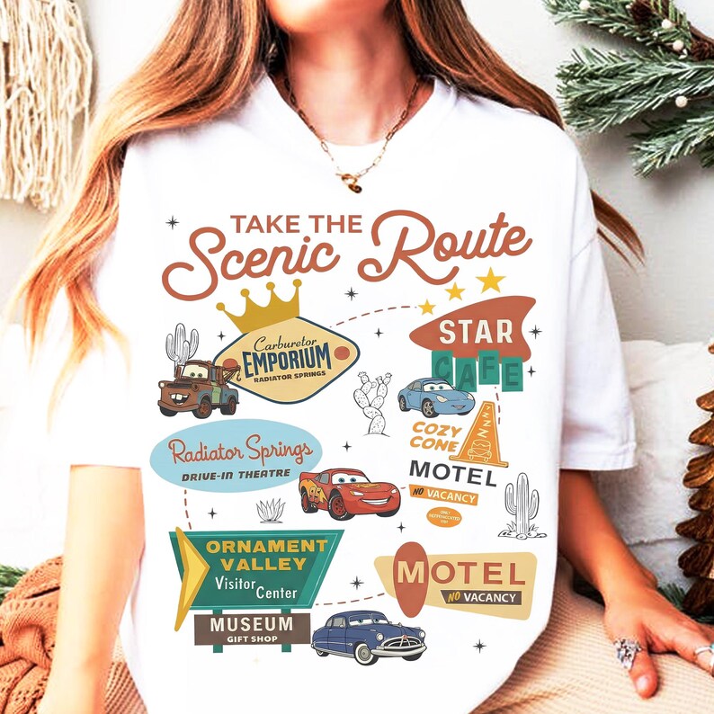 May include: White t-shirt featuring a colorful graphic design with the text "Take the Scenic Route." The design includes illustrations of cars, cacti, and various signs, including "Radiator Springs" and "Motel No Vacancy."