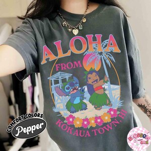 May include: Dark grey t-shirt with a colourful graphic reading "Aloha From Kokaua Town, HI." The design includes cartoon characters, a beach scene, and tropical flowers. The shirt has a vintage, faded appearance.
