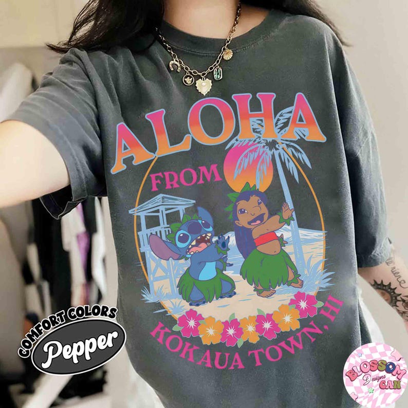 May include: Dark grey t-shirt with a colourful graphic reading "Aloha From Kokaua Town, HI." The design includes cartoon characters, a beach scene, and tropical flowers. The shirt has a vintage, faded appearance.