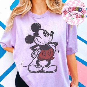Op de afbeelding: Een lichtpaars T-shirt met een geschetste Mickey Mouse-afbeelding in zwart, rood en wit. Het shirt heeft korte mouwen en een losse pasvorm. De afbeelding bevat de tekst "Blossom Designs Can" in een roze cirkel.