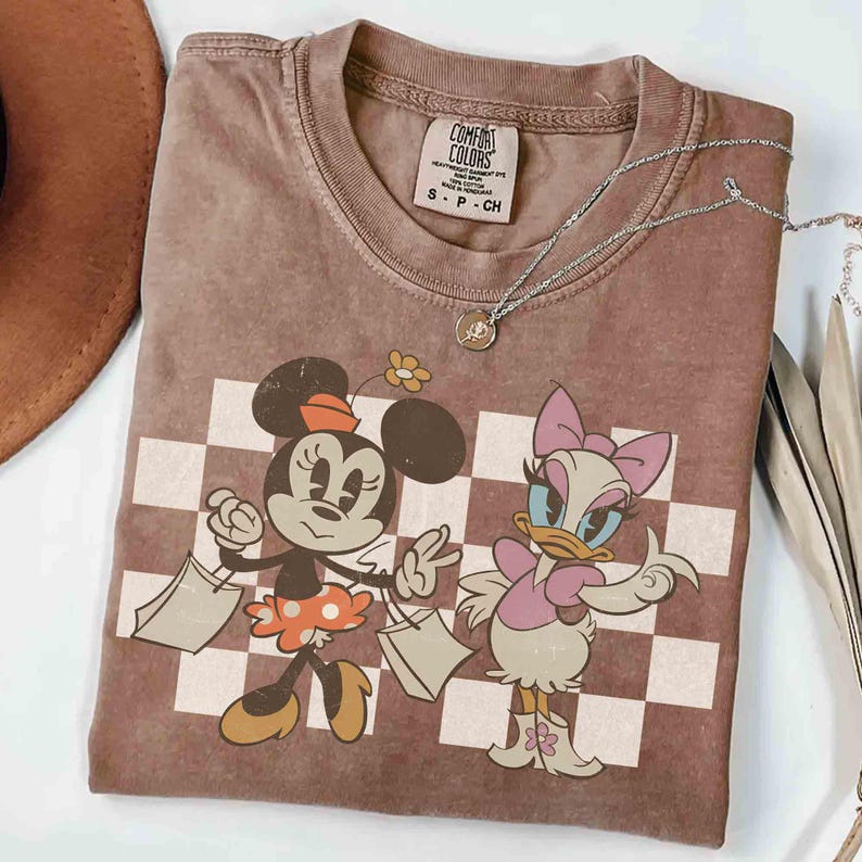 May include: A brown Comfort Colors t-shirt featuring Minnie Mouse and Daisy Duck with shopping bags, set against a checkered background. A silver necklace with a small pendant rests on the shirt. The shirt has a crew neck and short sleeves.