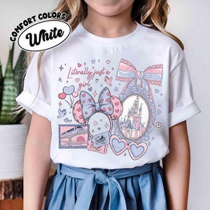 Puede incluir: Camiseta blanca Comfort Colors con un dise&ntilde;o gr&aacute;fico que presenta un castillo, gafas en forma de coraz&oacute;n y el texto "Literally just a girl". El dise&ntilde;o incluye lazos, corazones y un billete de tren.