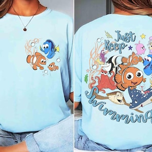 May include: Light blue t-shirt featuring cartoon characters from the movie "Finding Nemo". The front of the shirt shows Nemo, Dory, and a smaller fish. The back of the shirt has the phrase "Just Keep Swimming" with various sea creatures.