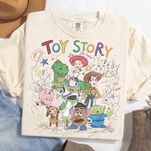 Puede incluir: Camiseta de color crema con un gr&aacute;fico colorido de los personajes de Toy Story, incluyendo a Woody, Buzz Lightyear y Rex. Las palabras "Toy Story" est&aacute;n escritas en colores arco&iacute;ris sobre los personajes. La camiseta tiene cuello redondo y mangas cortas.