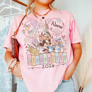 K&ouml;nnte beinhalten: Hellrosa T-Shirt mit einem farbenfrohen Disney-Motiv, darunter Micky Maus und Minnie Maus. Das Design enth&auml;lt den Text "Disney Trip 2026" und Platz f&uuml;r einen Namen.