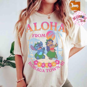 May include: Cream-coloured t-shirt with the words "Aloha From Kokaua Town, HI" in orange and pink. The design features cartoon characters Stitch and Lilo, palm trees, and flowers. The shirt has a relaxed fit.