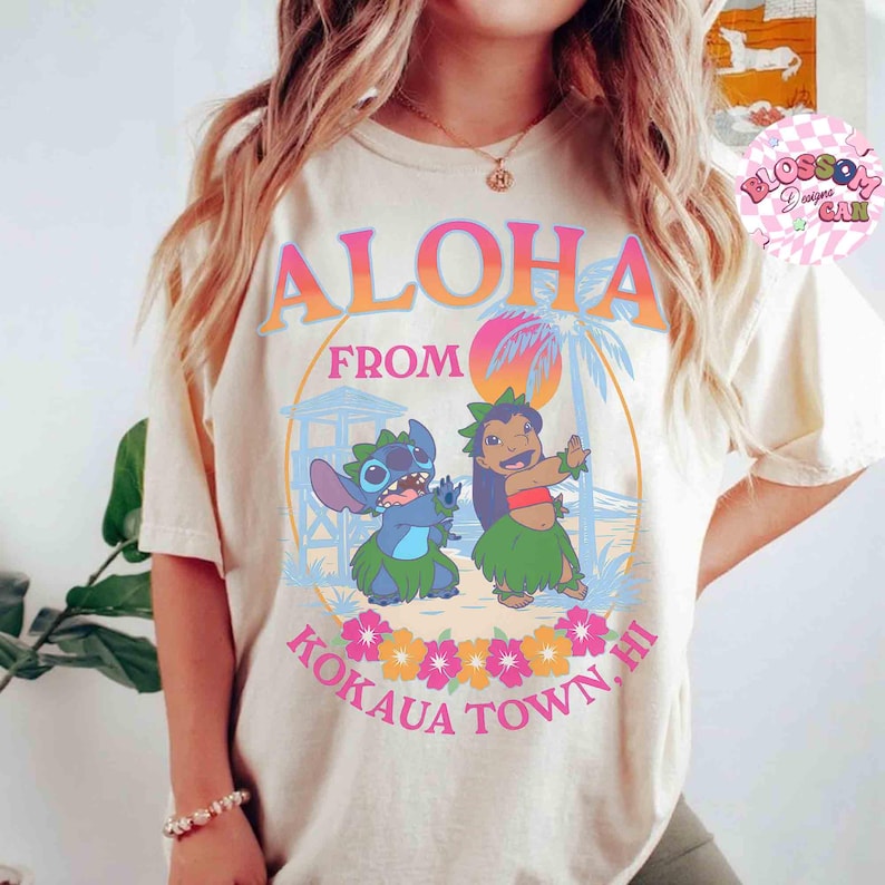 May include: Cream-coloured t-shirt with the words "Aloha From Kokaua Town, HI" in orange and pink. The design features cartoon characters Stitch and Lilo, palm trees, and flowers. The shirt has a relaxed fit.