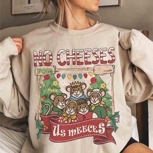 May include: Beige sweatshirt with a festive graphic. The design features a group of cartoon mice in front of Christmas trees, with the text "NO CHEESES FOR Us MEEECES" in a candy cane-style font. The design includes holly and string lights.