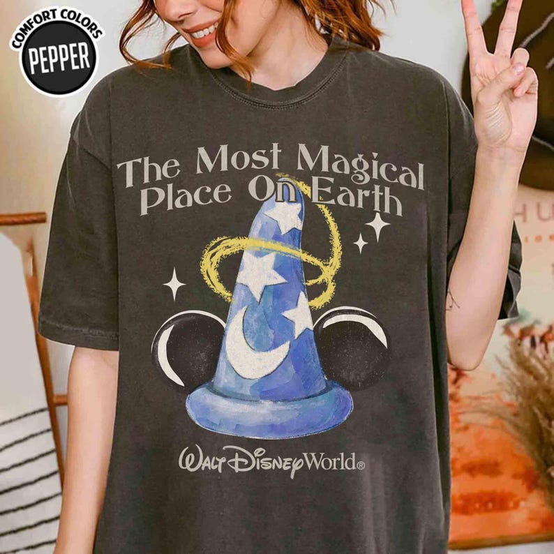 May include: Dark grey t-shirt with a blue wizard's hat design, white stars, and a crescent moon, encircled by a gold swirl. The text reads "The Most Magical Place On Earth" and "Walt Disney World".