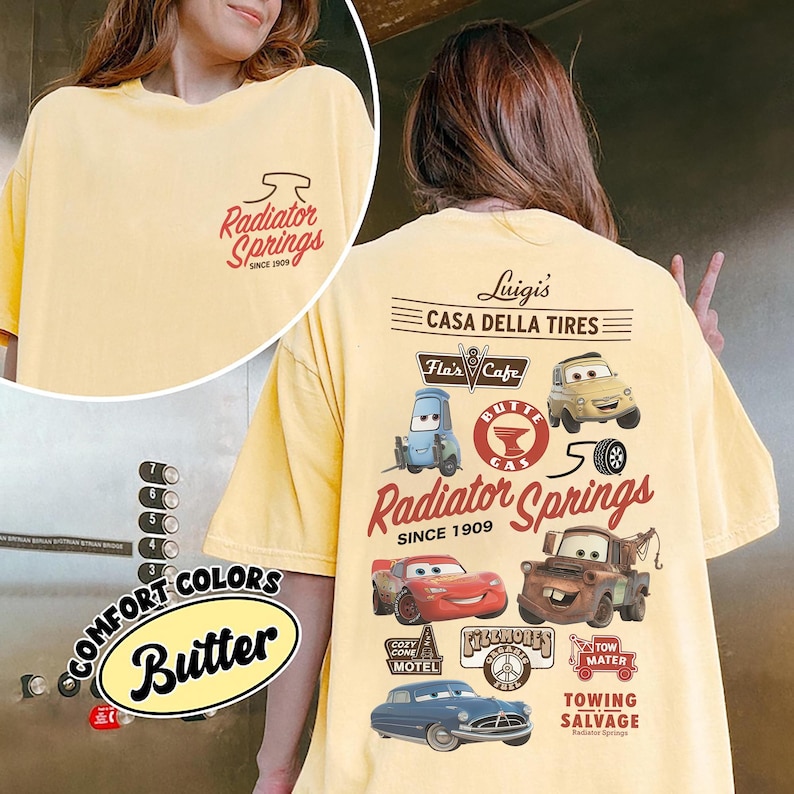 May include: A pale yellow t-shirt featuring a small Radiator Springs logo on the front chest. The back displays a large graphic with characters and locations from the movie Cars, including Luigi's Casa Della Tires, Flo's Cafe, and Mater.