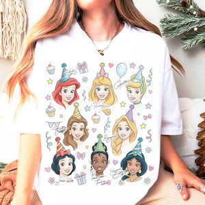 May include: White t-shirt featuring colorful illustrations of Disney princesses wearing party hats. The design includes cupcakes, stars, and the princesses' signatures. A festive and whimsical design.