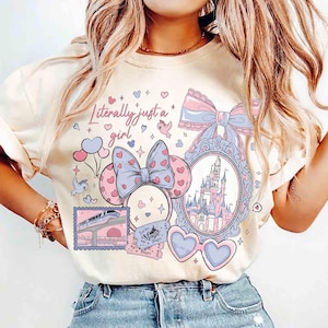 Puede incluir: Camiseta color crema con un dise&ntilde;o caprichoso que presenta un castillo, orejas de Minnie Mouse, gafas de sol en forma de coraz&oacute;n y la frase "Literally just a girl". El dise&ntilde;o incorpora elementos rosas, azules y morados.