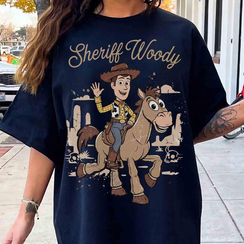 Op de afbeelding: Een donkerblauw t-shirt met een grafische print van Sheriff Woody op een bruin paard. De tekst "Sheriff Woody" staat in een goudkleurig cursief lettertype boven het personage. Het ontwerp bevat woestijncactussen en schedels.