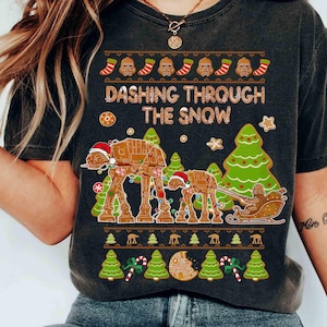 以下が含まれることがあります： 「Dashing Through The Snow」のデザインが施された、濃いグレーのTシャツ。サンタ帽をかぶったAT-ATウォーカー、そり、クリスマスツリー、ジンジャーブレッド風のアクセントが特徴です。楽しいホリデーテーマのTシャツ。