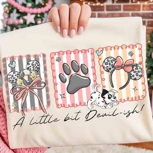 May include: Cream-colored t-shirt with three rectangular designs. The designs feature Dalmatian-themed mouse ears, a paw print, and a Dalmatian puppy. The text "A little bit Devil-ish!" is written in cursive at the bottom.