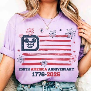 May include: A light purple t-shirt featuring a stylized American flag design with red stripes and a blue section incorporating a Mickey Mouse head. The shirt includes the text "250TH AMERICA ANNIVERSARY 1776-2026" in red and navy blue.