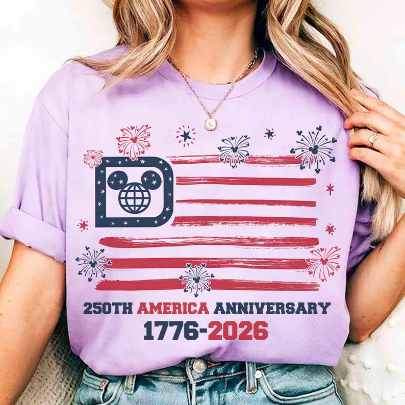 May include: A light purple t-shirt featuring a stylized American flag design with red stripes and a blue section incorporating a Mickey Mouse head. The shirt includes the text "250TH AMERICA ANNIVERSARY 1776-2026" in red and navy blue.