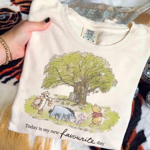 K&ouml;nnte beinhalten: Cremefarbenes Sweatshirt mit einer Grafik von Winnie Puuh und seinen Freunden unter einem gro&szlig;en Baum. Der Text "Today is my new favourite day" ist unter dem Bild aufgedruckt. Das Sweatshirt hat einen Rundhalsausschnitt und lange &Auml;rmel.