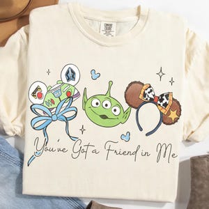 May include: Cream-colored t-shirt featuring a whimsical design with cartoon characters and the phrase "You've Got a Friend in Me." The design includes a Buzz Lightyear-themed bow, a green alien, and Mickey Mouse ears with a cow print.