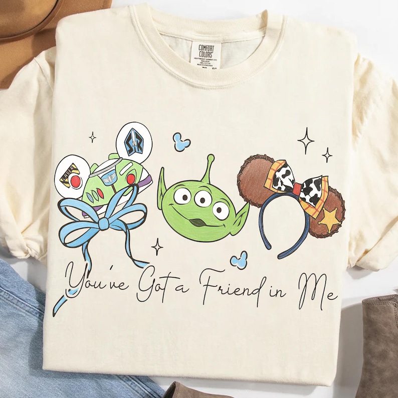 May include: Cream-colored t-shirt featuring a whimsical design with cartoon characters and the phrase "You've Got a Friend in Me." The design includes a Buzz Lightyear-themed bow, a green alien, and Mickey Mouse ears with a cow print.