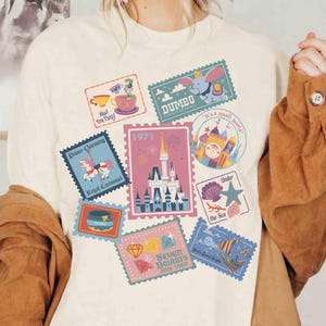 May include: A cream-colored t-shirt featuring a graphic print of various postage stamp-style illustrations. The designs include a castle, Dumbo, a carousel horse, a teacup, a ship, and text such as 1971, Prince Charming, and Seven Dwarfs Mine Train.