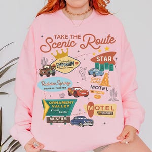 May include: Pink sweatshirt featuring a graphic design with the text "Take the Scenic Route" and various illustrations of cars, cacti, and signs. The design includes the words "Radiator Springs," "Motel," and "Museum."