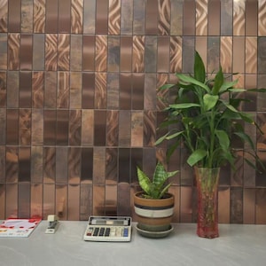 May include: A wall of copper-coloured tiles in a vertical rectangular design. A green plant in a clear vase and a smaller potted plant sit on a grey surface. A calculator, stapler, and magazine are also on the surface.